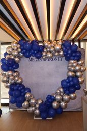 Circular balloon arch of royal blue and metallic silver balloons framing a gray backdrop in a modern indoor event space with wood floor and linear ceiling lights.