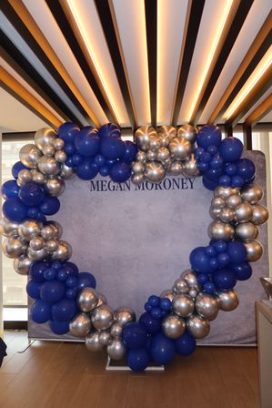 Circular balloon arch of royal blue and metallic silver balloons framing a gray backdrop in a modern indoor event space with wood floor and linear ceiling lights.