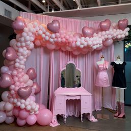 Pink party backdrop in an indoor event space with a pastel pink balloon arch and metallic heart balloons, vintage pink vanity mirror, two mannequins styled in pink and black dresses, and pink cowboy boots — feminine event decor and photo-booth display.