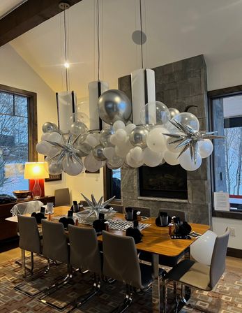 Modern dining room with a long wooden table set for eight under a hanging silver-and-white balloon chandelier with metallic starburst accents, stone fireplace and large windows showing snowy trees outside.
