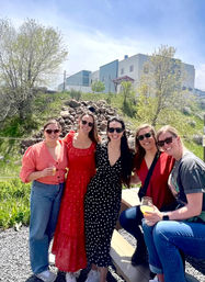 Group of five friends on a sunny outdoor patio holding drinks, wearing sunglasses and colorful outfits (red maxi dress, black polka-dot dress, jeans) with a grassy hillside and modern industrial-style building in the background