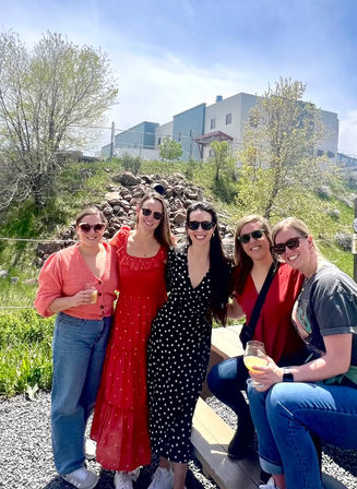Group of five friends on a sunny outdoor patio holding drinks, wearing sunglasses and colorful outfits (red maxi dress, black polka-dot dress, jeans) with a grassy hillside and modern industrial-style building in the background