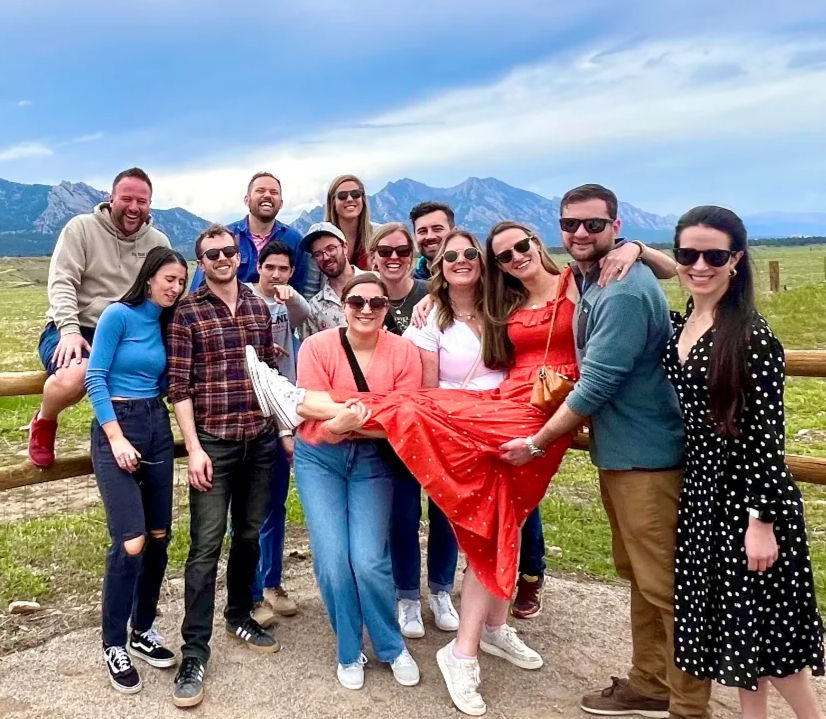 Smiling group of friends posing by a wooden fence in a grassy meadow, woman in a red dress playfully lifted, sunglasses and casual outfits, jagged mountain peaks and cloudy sky in the background.