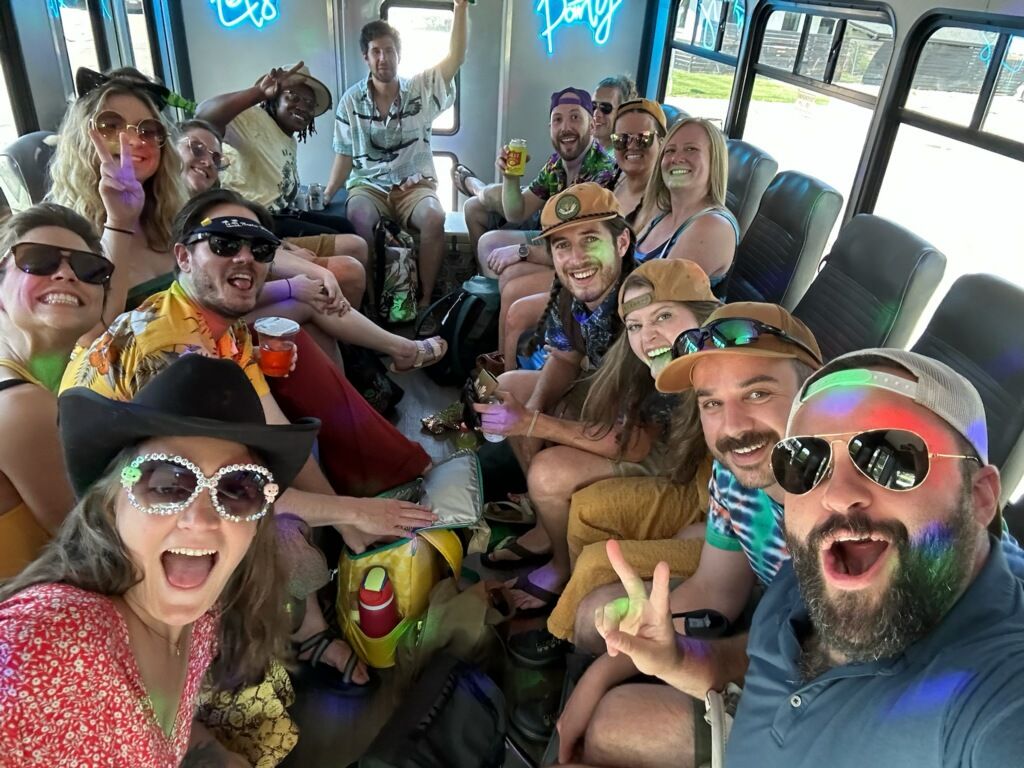 Excited group of friends on a daytime party bus, wearing sunglasses and hats, holding drinks and posing for a selfie amid colorful lights and a neon sign.