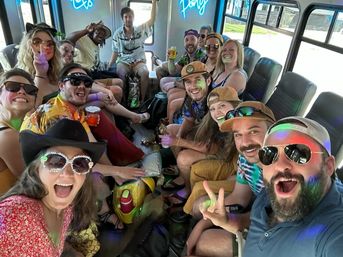 Excited group of friends on a daytime party bus, wearing sunglasses and hats, holding drinks and posing for a selfie amid colorful lights and a neon sign.