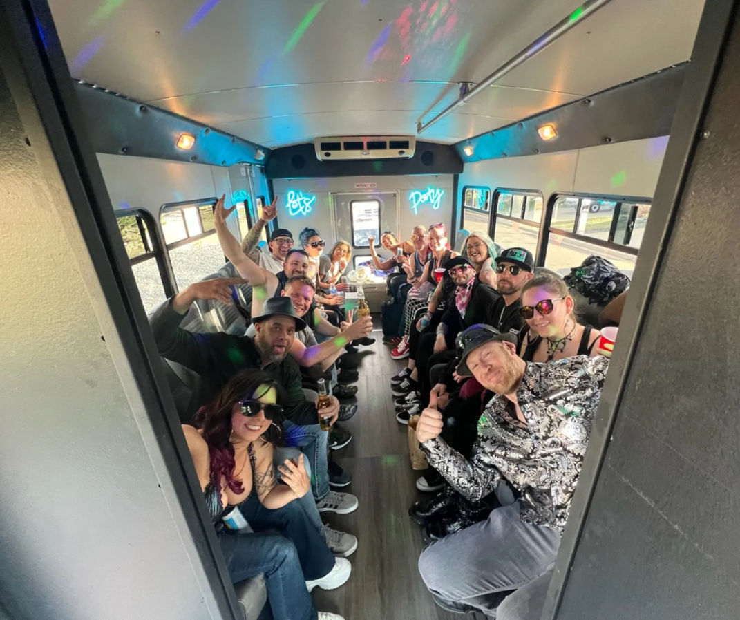 Lively group of people partying inside a party bus with bench seating, neon “Let’s Party” signs, colorful disco lights, drinks and thumbs-up.