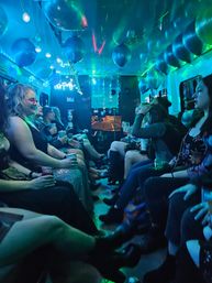 Neon-lit party bus interior at night with blue-green LED lights, hanging balloons, and a lively group seated along benches holding drinks — festive girls' night out vibe.