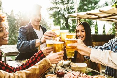 Friends clinking glasses of beer in a sunny outdoor patio toast over a picnic table with snacks and charcuterie, casual al fresco summer gathering