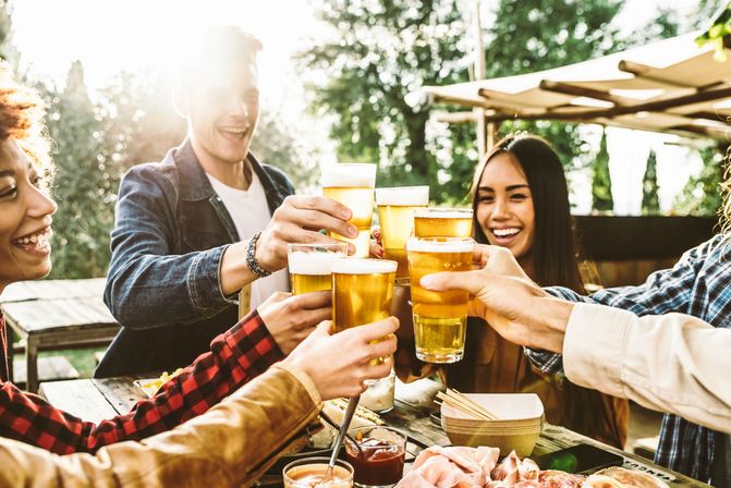 Friends clinking glasses of beer in a sunny outdoor patio toast over a picnic table with snacks and charcuterie, casual al fresco summer gathering