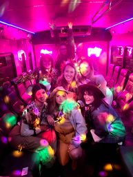 Group of friends wearing hats and playful accessories celebrating on a neon-pink party bus with rainbow disco lights and leather bench seating, lively nightlife vibe.