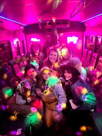 Group of friends wearing hats and playful accessories celebrating on a neon-pink party bus with rainbow disco lights and leather bench seating, lively nightlife vibe.