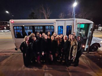 Smiling group of women on a city sidewalk at night posed in front of a white party shuttle with its door open — friends enjoying a night out.