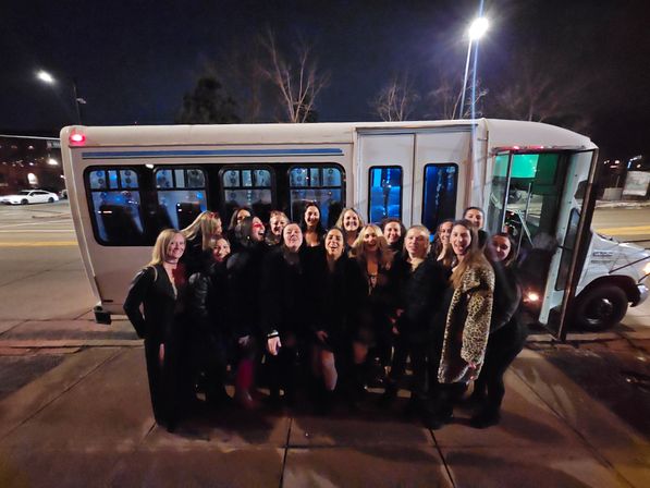 Smiling group of women on a city sidewalk at night posed in front of a white party shuttle with its door open — friends enjoying a night out.