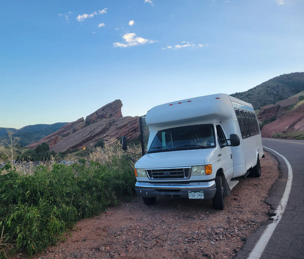 White passenger shuttle parked on a winding mountain road beside jagged red sandstone cliffs and green scrub under a bright blue sky — scenic western U.S. roadside.