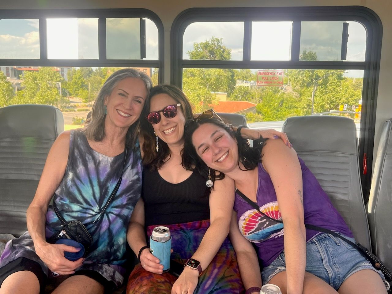 Three smiling friends in colorful tie-dye tops and sunglasses sitting close together on a daytime shuttle bus, holding canned drinks with green trees and buildings visible through the windows.