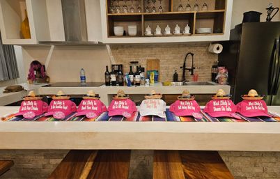 Party favors: row of pink and one white baseball caps topped with tiny straw sombreros, each reading “Most Likely To…”, arranged on a colorful Mexican serape across a modern kitchen island with bottles, glassware and a purple wig in the background.