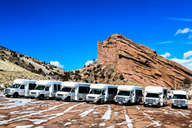 Row of white shuttle buses parked on a snowy lot in front of dramatic red sandstone cliffs under a vivid blue sky