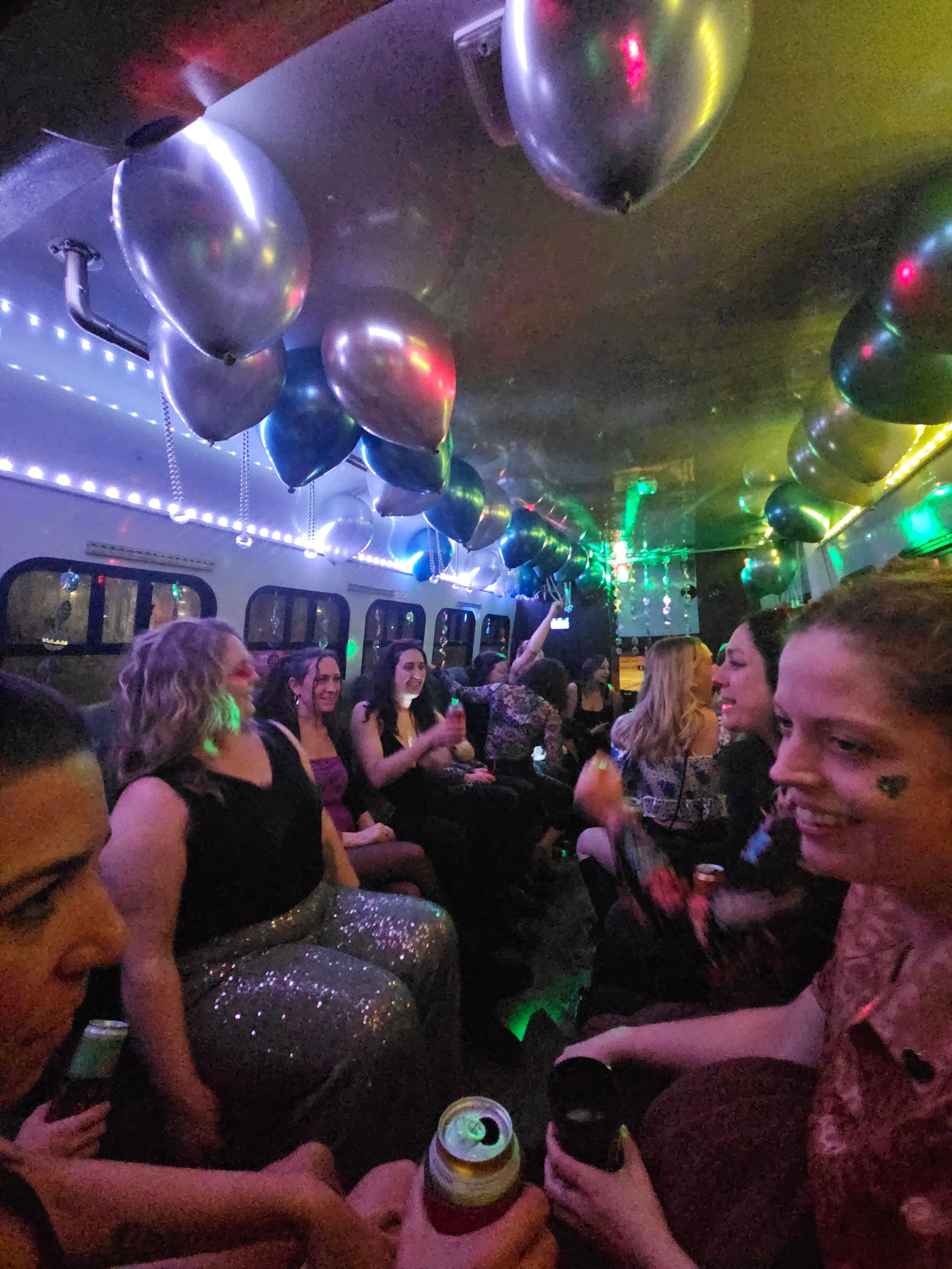 Party bus interior with a group of women celebrating under metallic balloons and colorful LED lights, people smiling and holding cans and cups in a festive nighttime scene