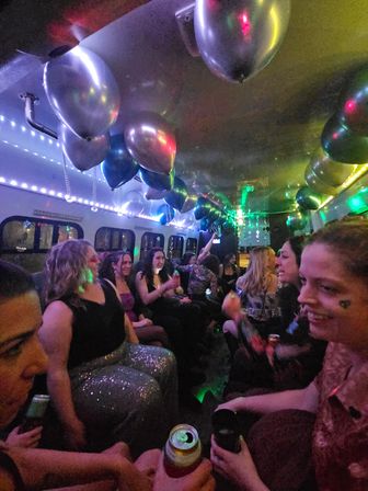 Party bus interior with a group of women celebrating under metallic balloons and colorful LED lights, people smiling and holding cans and cups in a festive nighttime scene