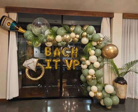 Indoor bachelorette party backdrop in a living room: sage green, white, and metallic gold balloon arch with oversized gold champagne bottle and ring balloons, gold foil letter balloons, and palm leaves framing sliding glass doors.