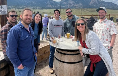 Smiling group of friends gathered around a wooden barrel table with drinks on an outdoor patio, with grassy valley and rocky mountain peaks in the scenic background.