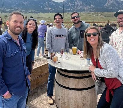 Smiling group of friends gathered around a wooden barrel table with drinks on an outdoor patio, with grassy valley and rocky mountain peaks in the scenic background.