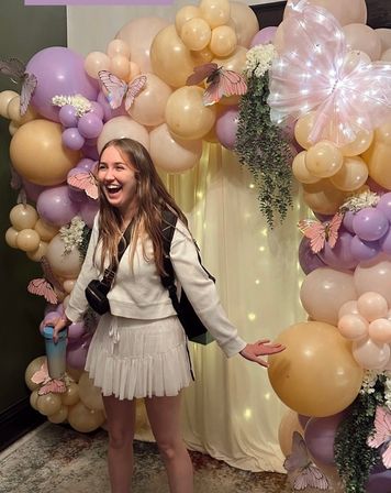 Smiling young woman posing in front of a pastel balloon arch and party backdrop with pink, lavender and gold balloons, butterfly decorations, greenery and fairy lights — indoor celebration scene.