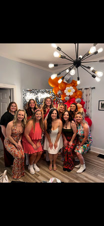 Group of women posing at an indoor celebration in a living room, smiling in front of a sequin photo wall and orange-pink balloon arch with disco balls and a daisy balloon, colorful retro dresses and a modern sputnik chandelier overhead.