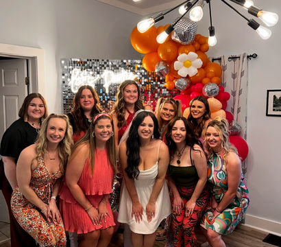 Group of women posing at an indoor celebration in a living room, smiling in front of a sequin photo wall and orange-pink balloon arch with disco balls and a daisy balloon, colorful retro dresses and a modern sputnik chandelier overhead.