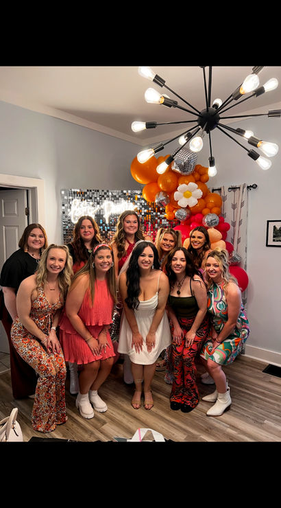 Group of women posing at an indoor celebration in a living room, smiling in front of a sequin photo wall and orange-pink balloon arch with disco balls and a daisy balloon, colorful retro dresses and a modern sputnik chandelier overhead.