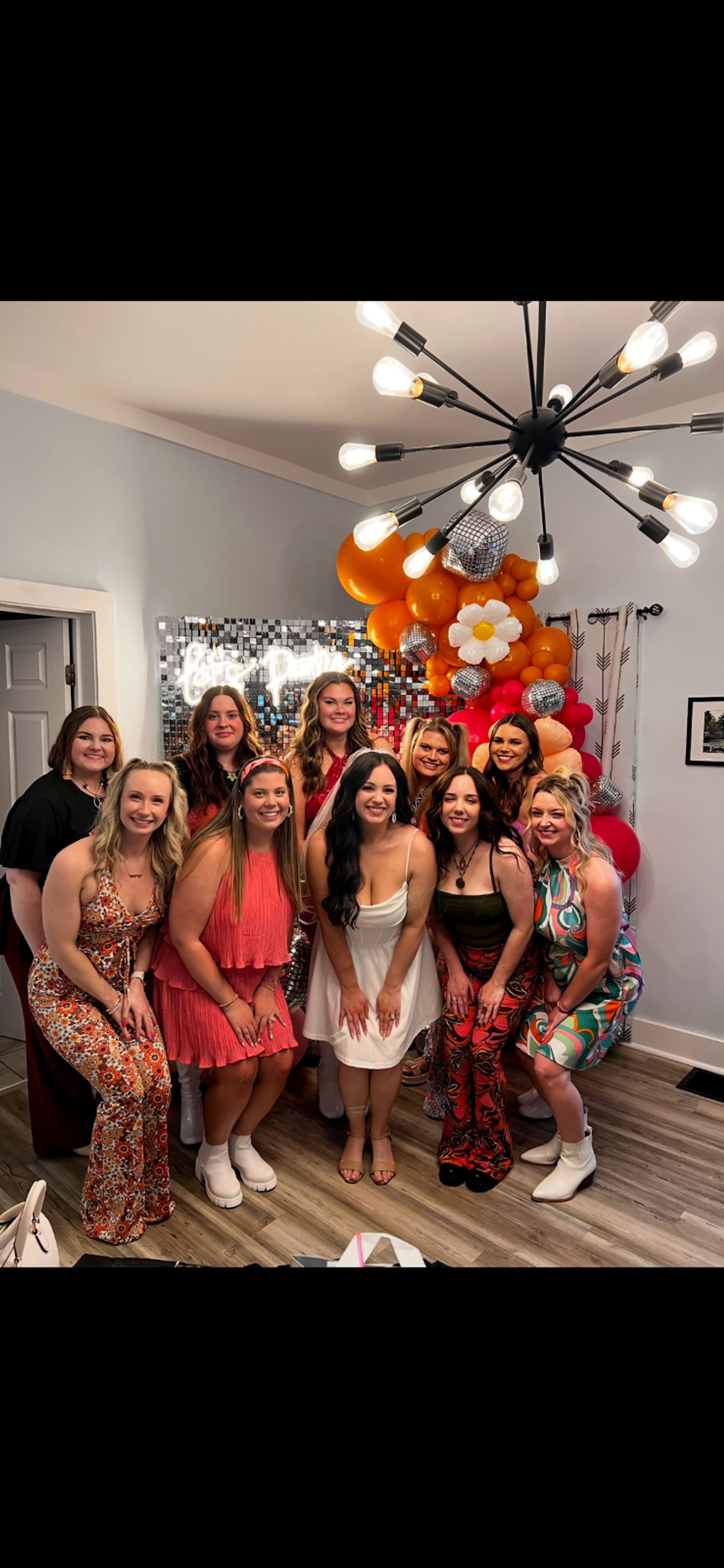 Group of women posing at an indoor celebration in a living room, smiling in front of a sequin photo wall and orange-pink balloon arch with disco balls and a daisy balloon, colorful retro dresses and a modern sputnik chandelier overhead.
