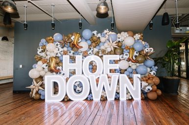 Illuminated HOEDOWN letters in a rustic indoor loft, fronting a western-themed balloon wall of blue, tan and cow-print balloons with gold cowboy boot and star accents on polished hardwood floors.