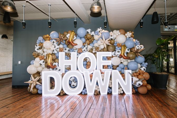 Illuminated HOEDOWN letters in a rustic indoor loft, fronting a western-themed balloon wall of blue, tan and cow-print balloons with gold cowboy boot and star accents on polished hardwood floors.