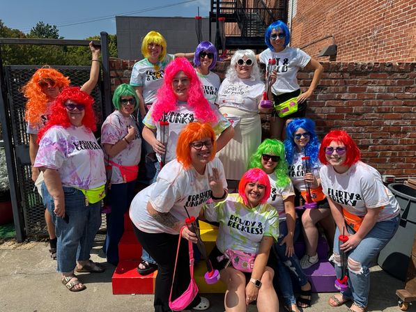 Group of women in bright colored wigs and sunglasses wearing matching tie-dye and slogan T-shirts, neon fanny packs and holding novelty cup plungers, posing on rainbow steps against a red brick urban patio wall