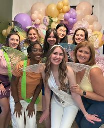 Happy friends in fairy wings pose with a bride-to-be sash at an indoor pastel balloon and butterfly-themed bridal shower.