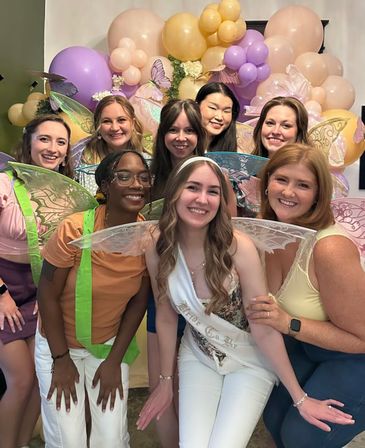 Happy friends in fairy wings pose with a bride-to-be sash at an indoor pastel balloon and butterfly-themed bridal shower.