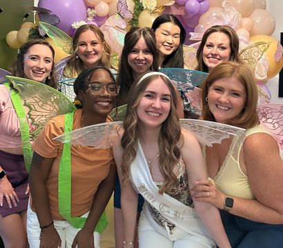 Happy friends in fairy wings pose with a bride-to-be sash at an indoor pastel balloon and butterfly-themed bridal shower.