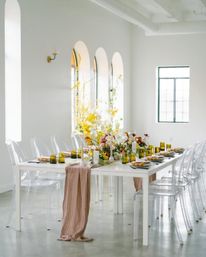 Stylish event table in a bright airy loft — white table with clear acrylic chairs, blush runner, yellow glassware and a lush floral centerpiece beneath arched windows.