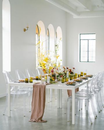 Stylish event table in a bright airy loft — white table with clear acrylic chairs, blush runner, yellow glassware and a lush floral centerpiece beneath arched windows.