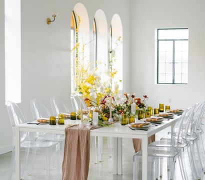 Stylish event table in a bright airy loft — white table with clear acrylic chairs, blush runner, yellow glassware and a lush floral centerpiece beneath arched windows.