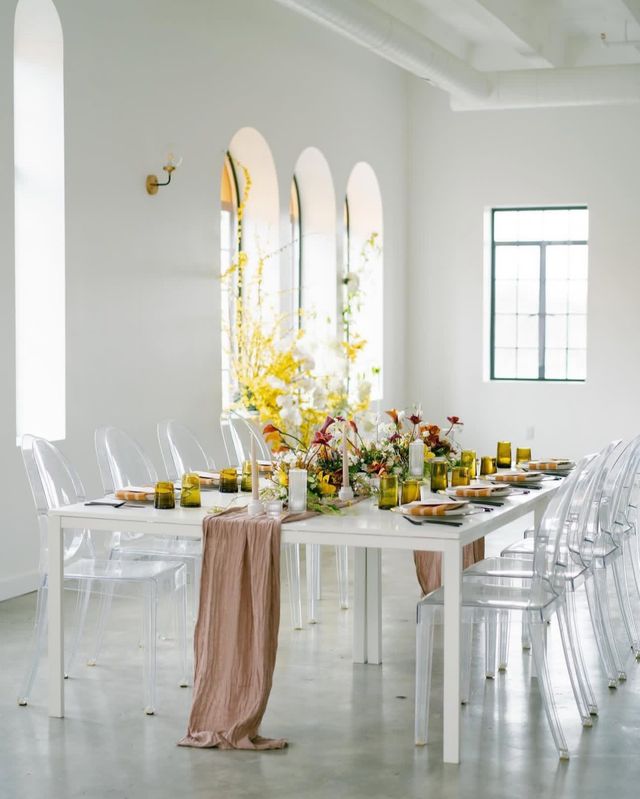 Stylish event table in a bright airy loft — white table with clear acrylic chairs, blush runner, yellow glassware and a lush floral centerpiece beneath arched windows.