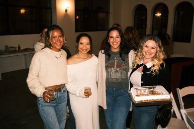 Four smiling women at an indoor evening party in a cozy event space with arched windows — two holding drinks and one carrying a cake, wearing casual sweaters and jeans.