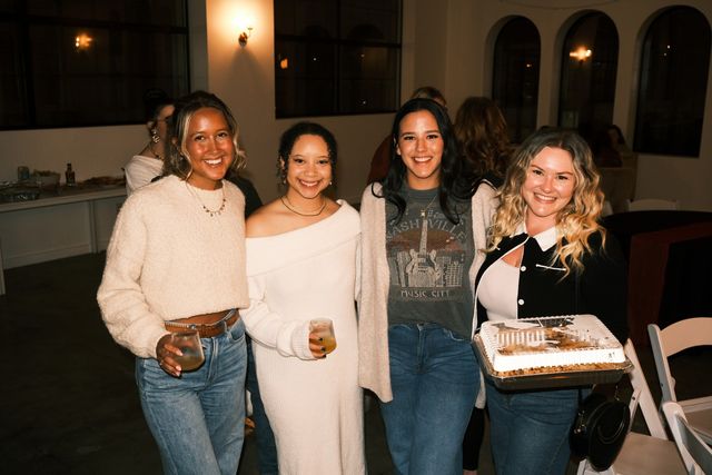 Four smiling women at an indoor evening party in a cozy event space with arched windows — two holding drinks and one carrying a cake, wearing casual sweaters and jeans.