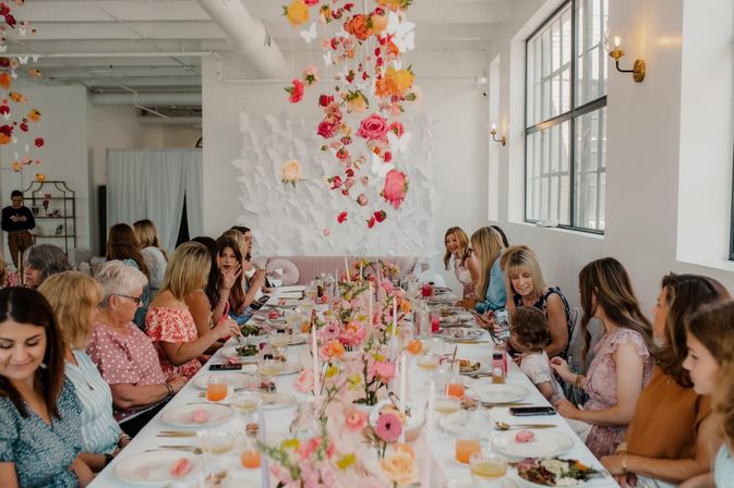 Pastel floral brunch in a bright urban loft — women gathered at a long table decorated with hanging roses, paper butterflies, and pink centerpieces.