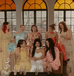 Group of laughing women in colorful dresses gathered on a couch in a sunlit room with arched amber windows and iced coffees on a low table, celebratory vibe.