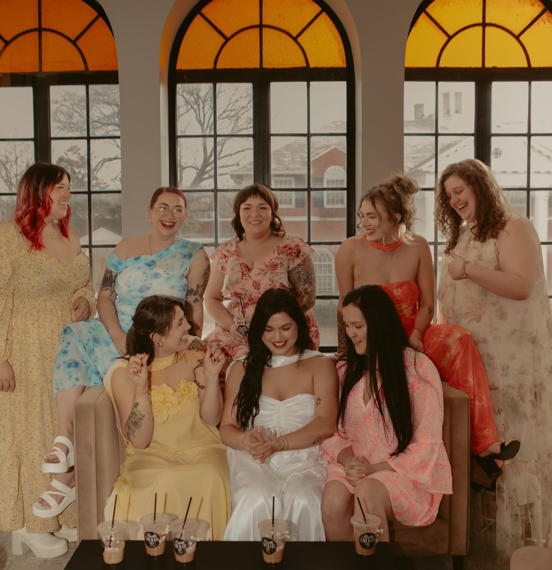Group of laughing women in colorful dresses gathered on a couch in a sunlit room with arched amber windows and iced coffees on a low table, celebratory vibe.