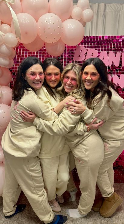 Four friends in matching cream loungewear and pink-tinted sunglasses hugging in front of a pink sequin backdrop and pale pink balloon arch decorated with pearls and ribbons at an indoor celebration.