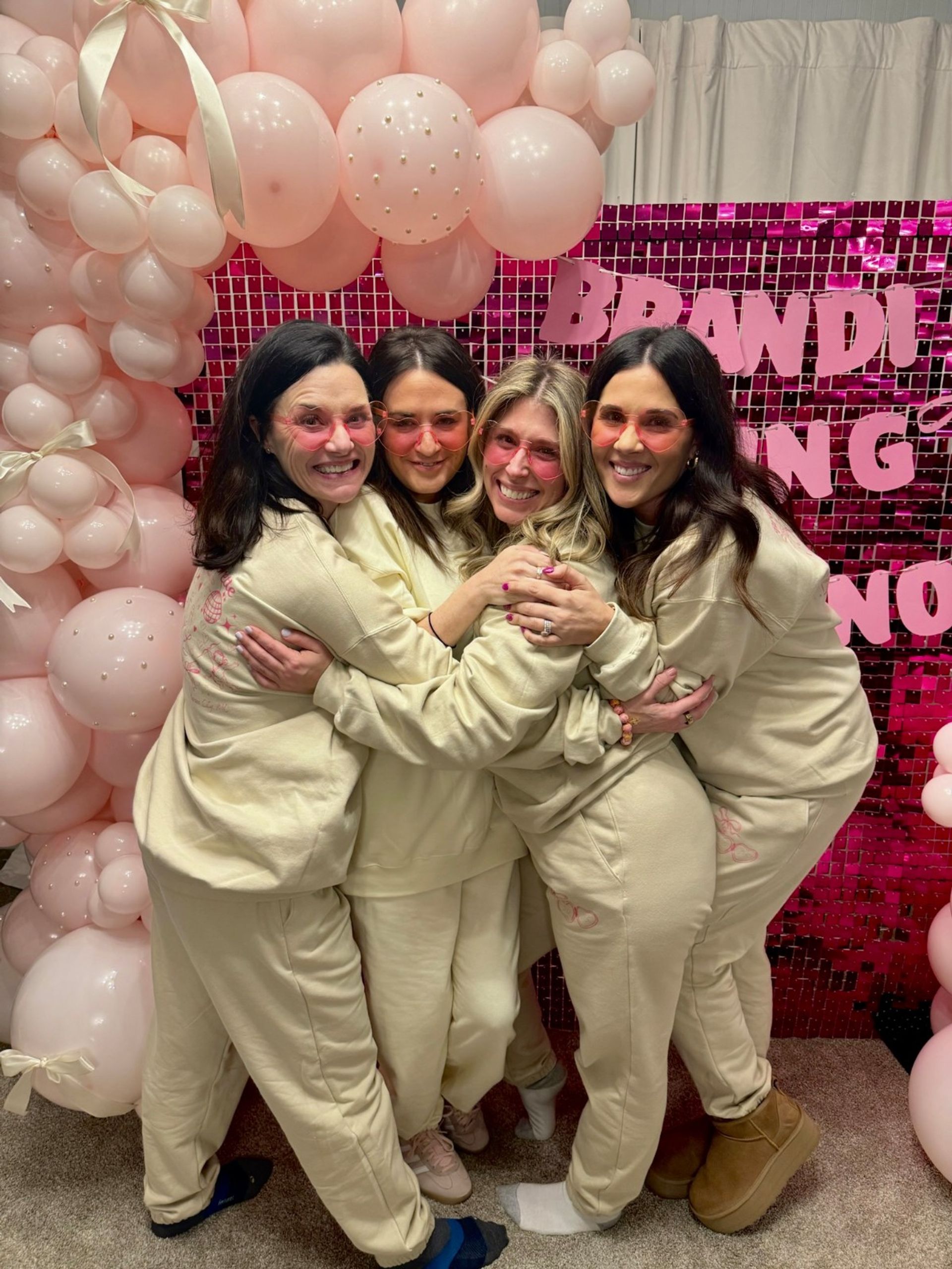 Four friends in matching cream loungewear and pink-tinted sunglasses hugging in front of a pink sequin backdrop and pale pink balloon arch decorated with pearls and ribbons at an indoor celebration.
