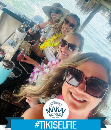 Four friends in sunglasses and colorful leis smiling for a selfie at a tiki-style boat bar over turquoise ocean while holding tropical drinks.