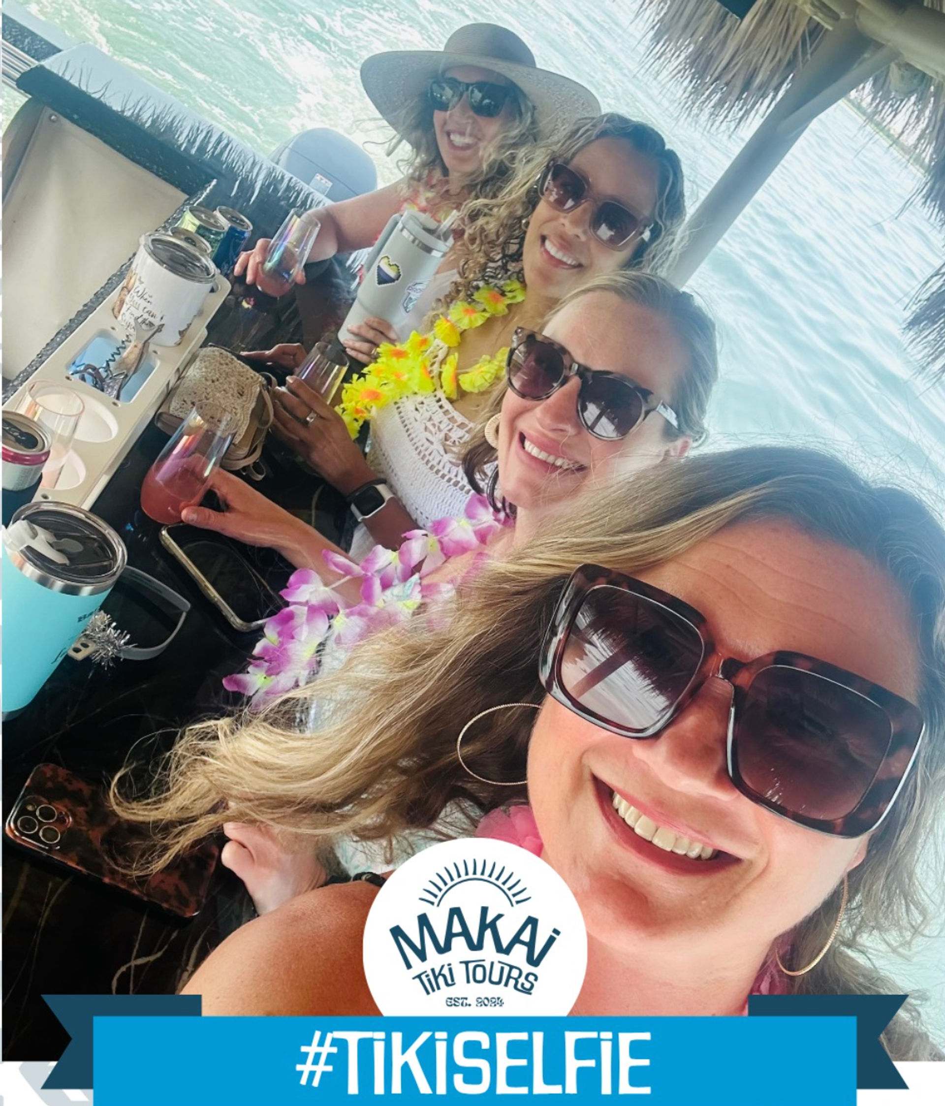 Four friends in sunglasses and colorful leis smiling for a selfie at a tiki-style boat bar over turquoise ocean while holding tropical drinks.